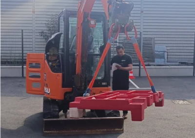 Un homme en tenue de sécurité se tient à côté d'un mini-excavateur orange avec un bras de levage rouge, dans une zone industrielle