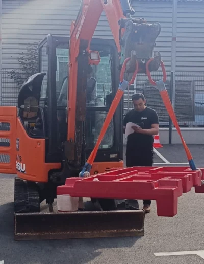 Un homme en tenue de sécurité se tient à côté d'un mini-excavateur orange avec un bras de levage rouge, dans une zone industrielle