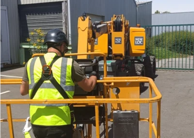 Un homme en vêtements de sécurité jaune et casque noir manipule les commandes d'une nacelle élévatrice jaune de la marque NEWLOC, dans une zone industrielle