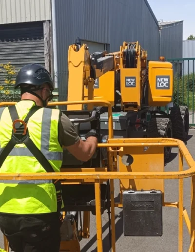 Un homme en vêtements de sécurité jaune et casque noir manipule les commandes d'une nacelle élévatrice jaune de la marque NEWLOC, dans une zone industrielle