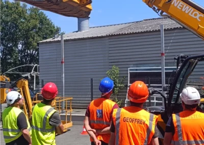 Un groupe d'hommes en gilets de sécurité orange et jaunes, portant des casques de protection, observent un engin de chantier et TP en action.