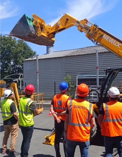Un groupe d'hommes en gilets de sécurité orange et jaunes, portant des casques de protection, observent un engin de chantier et TP en action.